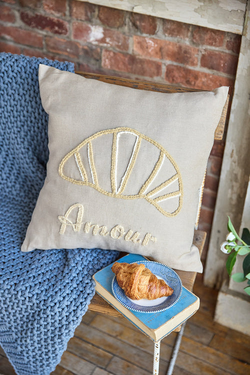 Beige square pillow with an embroidered croissant and the word 'amour'.