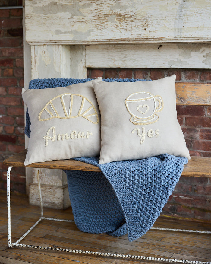 Beige square pillow with an embroidered croissant and the word 'amour'. Beige square pillow with an embroidered coffee cup and the word 'yes'.