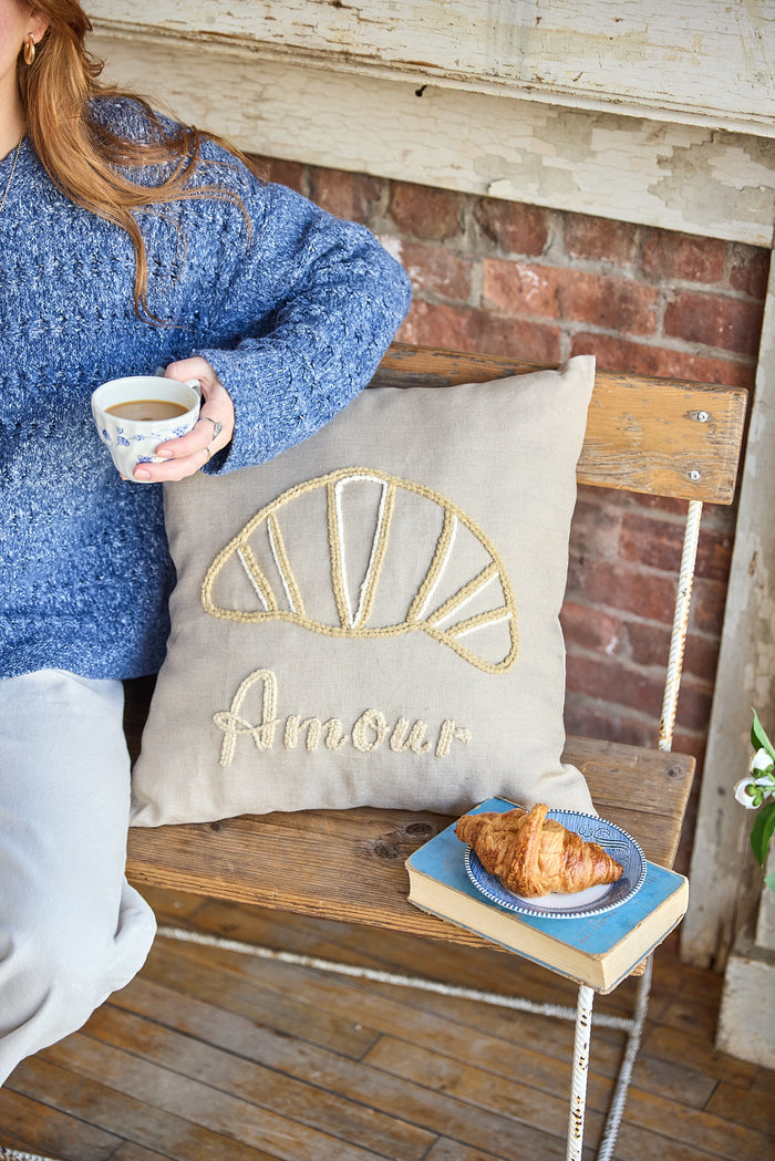 Person sitting next to a beige square pillow with an embroidered croissant and the word 'amour'.