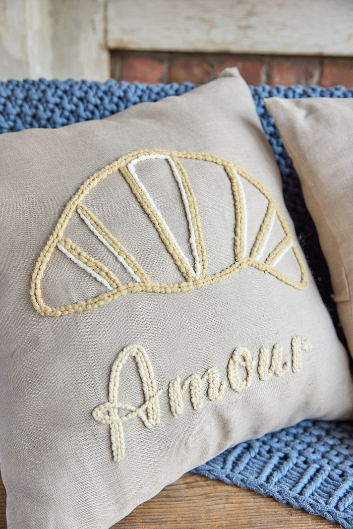 Beige square pillow with an embroidered croissant and the word 'amour'.
