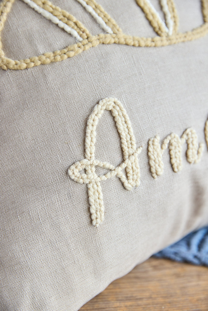 Beige square pillow with an embroidered croissant and the word 'amour'.