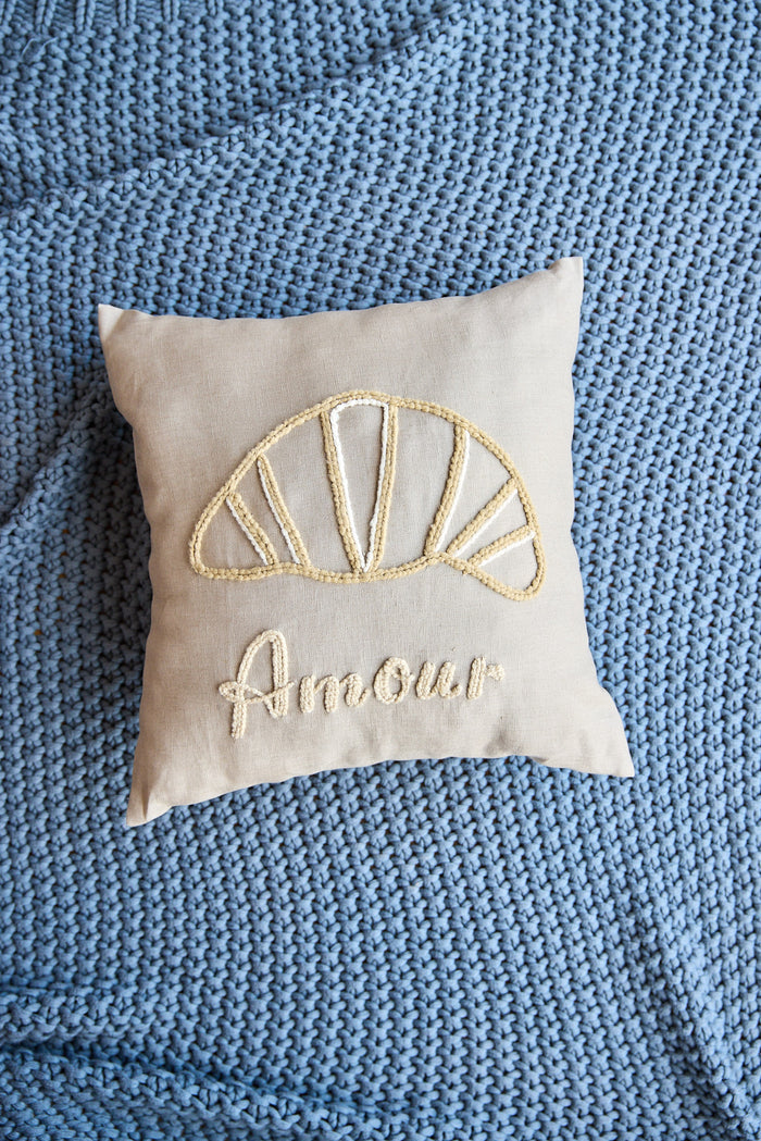 Beige square pillow with an embroidered croissant and the word 'amour'.