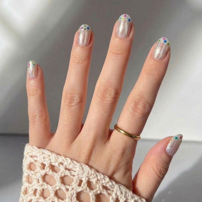 A hand wears a gold ring and a cream crochet sleeve. The Birthday Cake Press-On Nails by EyeCandys feature shimmery polish with playful, colorful flower designs near the tips for a fun, festive look.