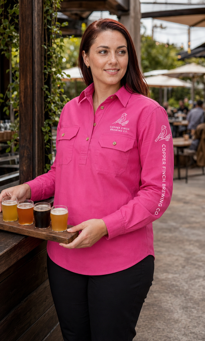 A woman in a bright pink long-sleeve button-up shirt stands at an outdoor bar holding a wooden tray with four beer glasses. The shirt has white printed logos on the chest and sleeve, including 'Copper Ridge Brewing Co.