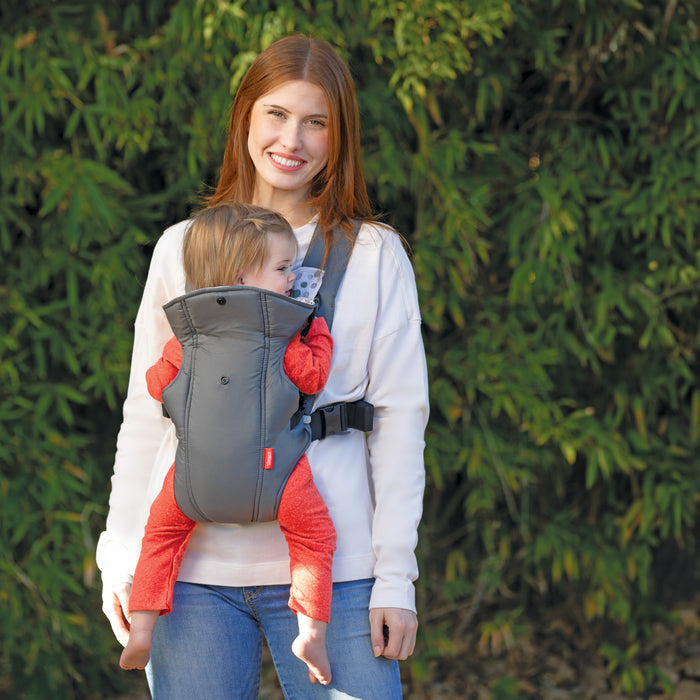 A smiling woman with long red hair wears a white sweater and blue jeans while carrying a toddler in the Nuby Classic Baby Carrier | Grey with breathable mesh, outdoors among greenery. Suitable for newborns to toddlers.