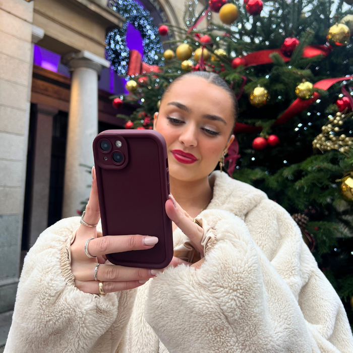 Woman in a beige coat takes a selfie holding a burgundy soft-finish phone case near a decorated Christmas tree.