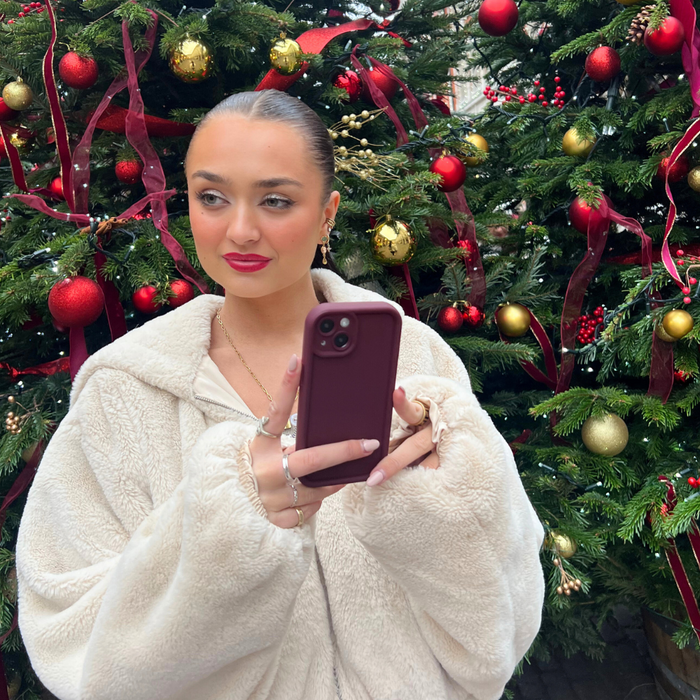 Woman in a white faux fur coat takes a selfie using a pale Cloud Soft NAKD phone case by Coconut Lane near Christmas trees.