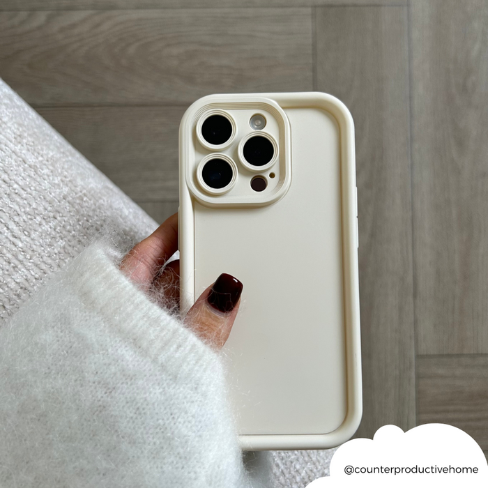 Hand with dark nails holds a cream Cloud Soft NAKD phone case by Coconut Lane, shown against a wood floor.