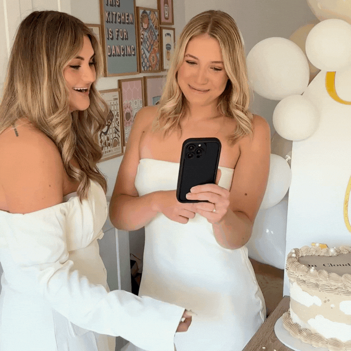 Two women in white dresses take a selfie at a party, holding a black Coconut Lane Cloud Soft NAKD phone case.