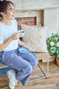 A person sitting on a chair next to a beige square pillow with an embroidered coffee cup and the word 'yes'.