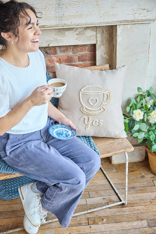 A person sitting on a chair next to a beige square pillow with an embroidered coffee cup and the word 'yes'.