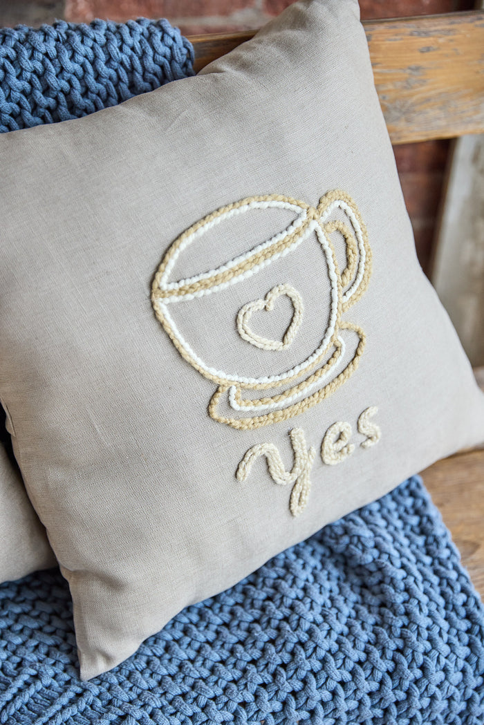 Beige square pillow with an embroidered coffee cup and the word 'yes'.