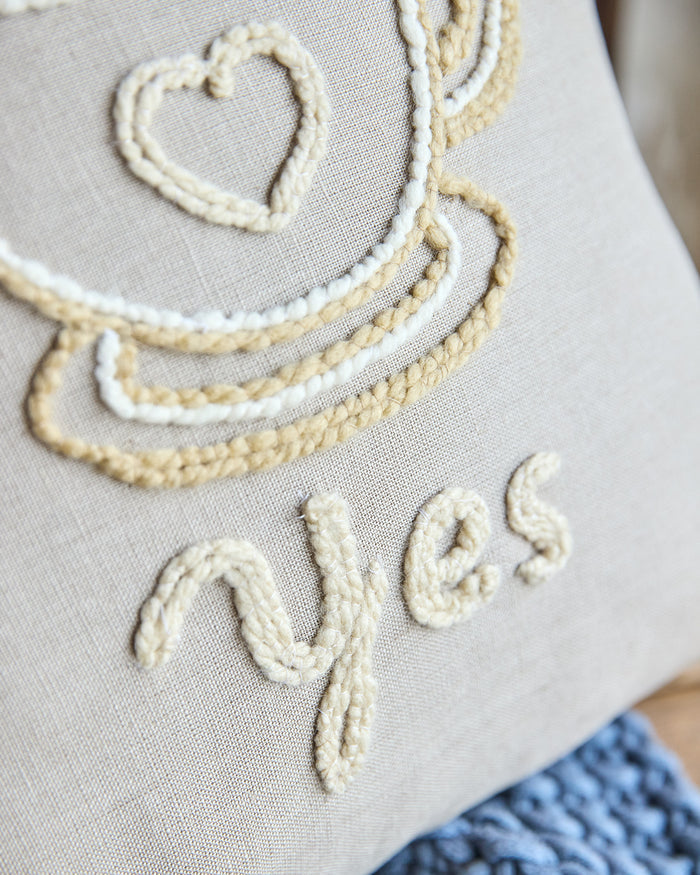 Beige square pillow with an embroidered coffee cup and the word 'yes'.