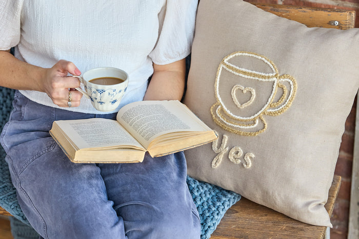 A person sitting next to a beige square pillow with an embroidered coffee cup and the word 'yes'.
