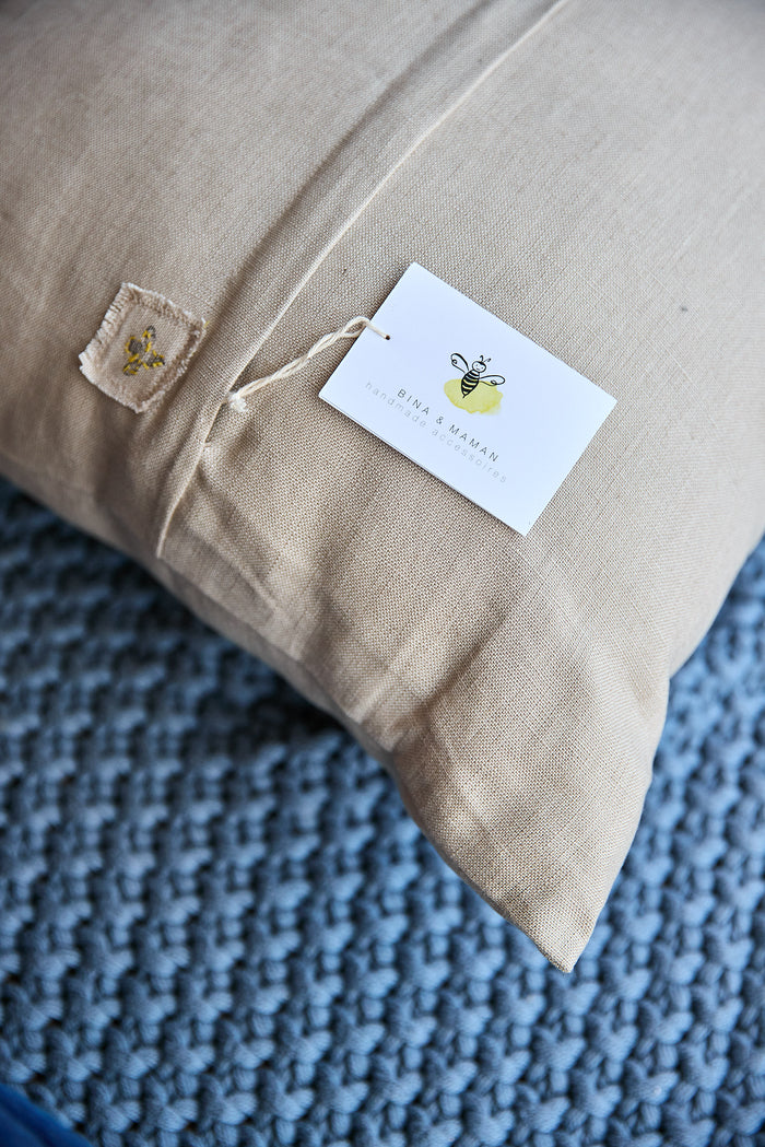 Back corner of a beige square pillow showing the 'bina x maman' label.