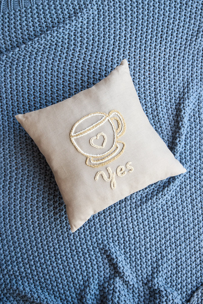 Beige square pillow with an embroidered coffee cup and the word 'yes'.