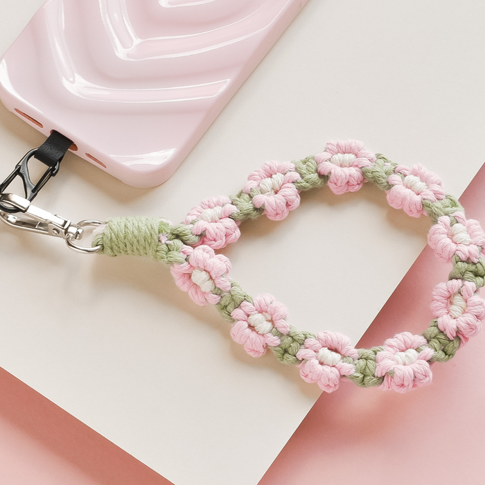 Pink phone with a pink and green crochet flower strap, displayed on a pink and white background.