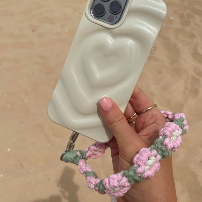 Hand holding a phone with a pink and green crochet flower strap, above sandy ground.