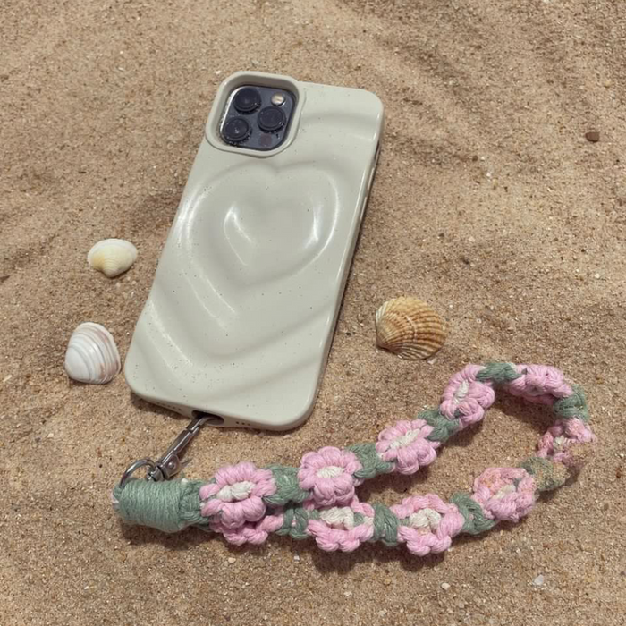 Pink and green crochet flower phone straps beside a heart phone case decorated with seashells on sand.