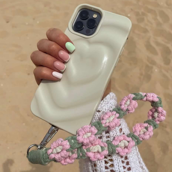 Hand with pastel pink and green nails holding a phone with a pink and green crochet flower phone strap attached.