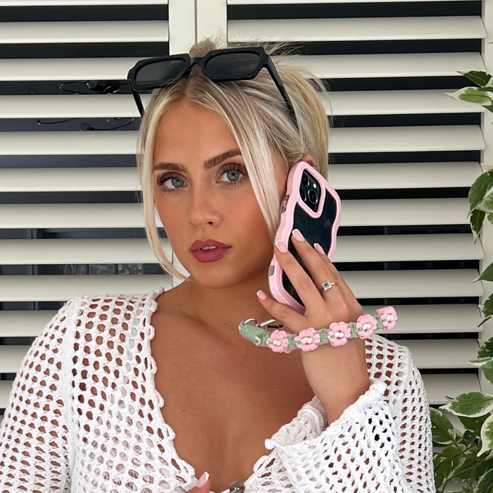 Blonde woman in sunglasses holds a pink Coconut Lane Curvy phone case to her ear, standing by white blinds.