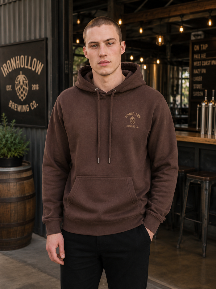 A man wearing a brown pullover hoodie with a drawstring hood and a kangaroo pocket, standing in a brewery setting. The hoodie features a small embroidered 'Ironhollow Brewing Co.