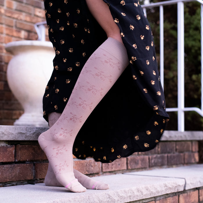 Knee-high compression socks with a floral texture, worn with a long black dress, resting on a stone step.