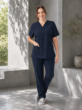 A woman wearing a matching navy blue scrub set with a V-neck top and straight-leg pants, standing in a modern concrete walkway with soft shadows.