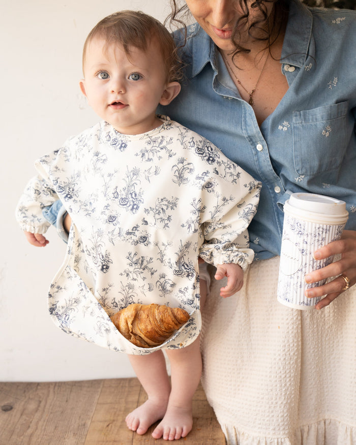 Baby wearing a blue floral toile bib with sleeves, with a croissant in the pocket.