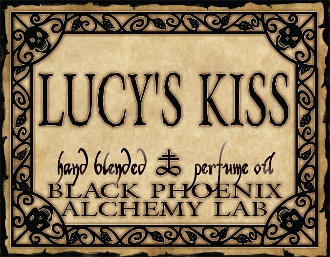 Label for 'Lucy's Kiss' hand blended perfume oil by Black Phoenix Alchemy Lab with decorative border.