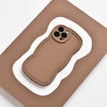 A lightweight, brown NAKD Curvy Phone Case - Coffee from Coconut Lane with a wavy design lies on a flat, brown and white, wavy-edged surface. The case has precise cutouts for the camera lenses and appears highly protective. The background contrasts with the NAKD Curvy Phone Case, highlighting its unique shape and design.