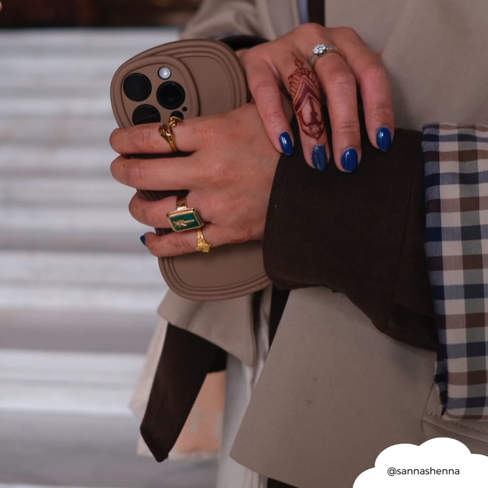 Blue-nailed hand with gold rings holds a phone in a brown NAKD Curvy Phone Case by Coconut Lane.