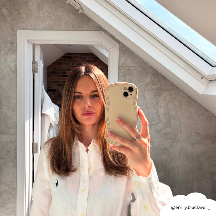 Woman with straight brown hair takes a mirror selfie in a bright bathroom, holding a vanilla-colored curvy phone case.