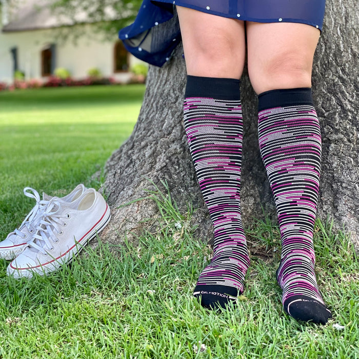Knee-high compression socks with a multicolored striped pattern, worn by a person sitting against a tree.