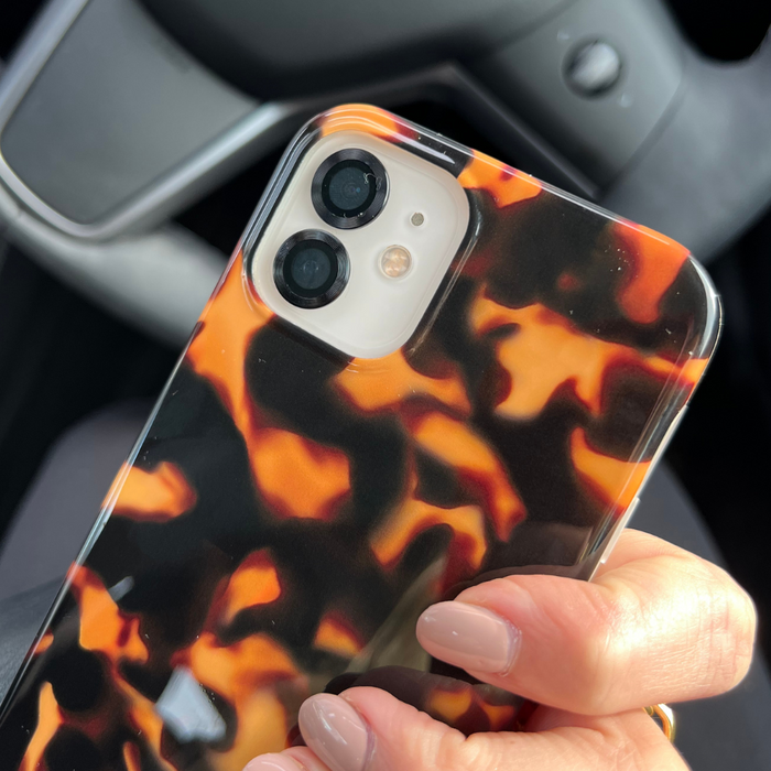 Hand holding an iPhone with a glossy tortoiseshell case and black camera lens covers inside a car.