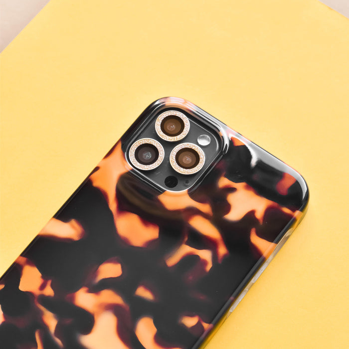 Smartphone in a black and orange tortoiseshell case with gold glitter camera lens covers on a yellow background.