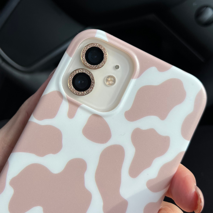 Phone held in hand with a pink and white cow-print case, rhinestones, and a gold glitter camera lens cover.