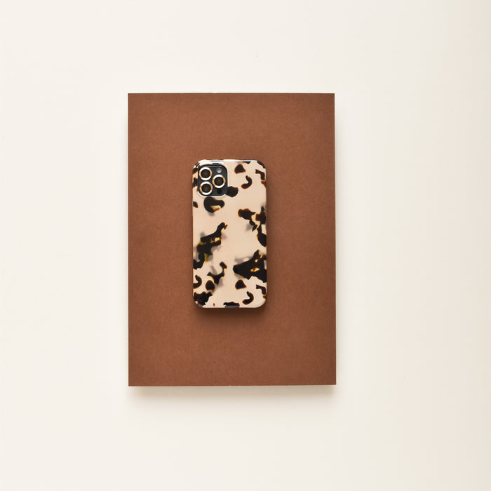 Smartphone in a brown and black tortoiseshell case with gold Coconut Lane camera lens covers, on a brown surface.