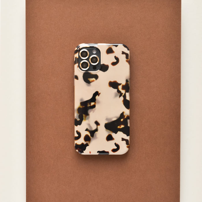 IPhone in a brown-black tortoiseshell case with gold Coconut Lane camera lens covers on a brown surface.