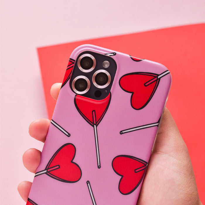 Hand holding a phone with a pink case, red heart-shaped lollipops, and pink Coconut Lane camera lens covers.