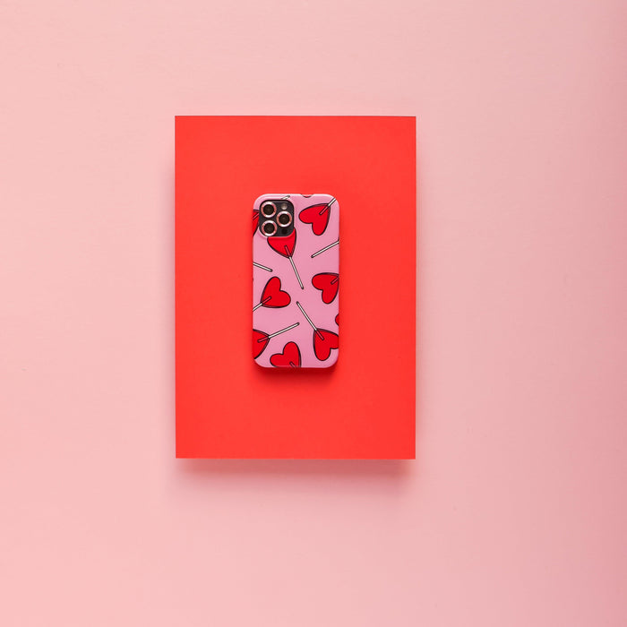 Smartphone with a pink heart case and pink Coconut Lane camera lens covers on a red rectangle, against a pink background.