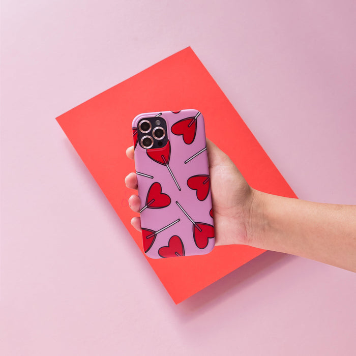 Hand holding a phone with pink Coconut Lane camera lens covers, shown over red paper on a pink background.
