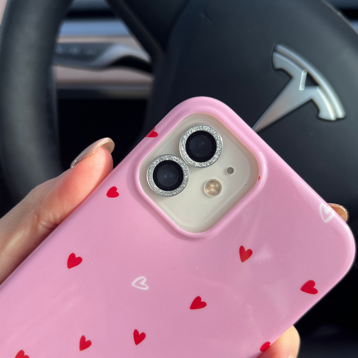 Pink phone with red hearts and a silver glitter camera lens cover held in front of a Tesla wheel.