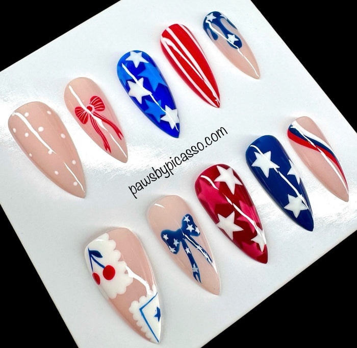 Ten press-on nails with patriotic red, white, and blue designs, including stars, stripes, and bows, displayed on a white card with the text 'pawsbypicaso.com'.