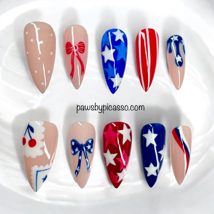 Ten almond-shaped press-on nails arranged in two rows on a white surface. The nails feature patriotic red, white, and blue designs, including stars, stripes, bows, and cherries, with a watermark 'pawsbypicasso.