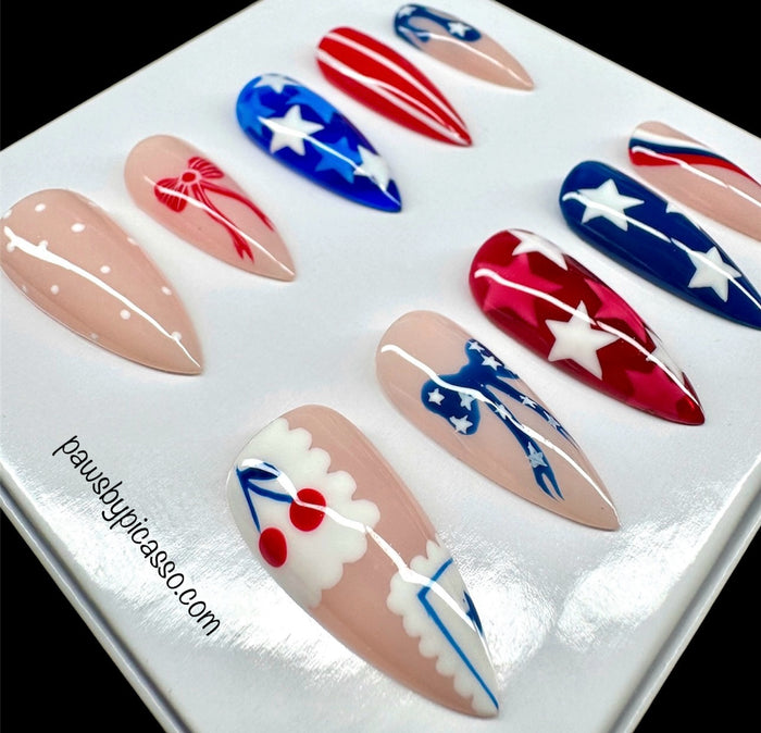 A set of press-on false nails displayed on a white card, featuring patriotic red, white, and blue designs including stars, stripes, bows, cherries, and polka dots on nude-toned bases.