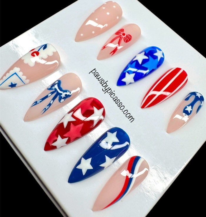 A set of 10 press-on nails displayed on a white backing card. The nails feature patriotic red, white, and blue designs, including stars, stripes, and bows, with some nails showing a nude base.