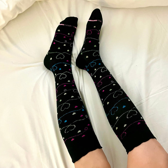 Knee-high black compression socks with colorful heart patterns, resting on a white bedspread.