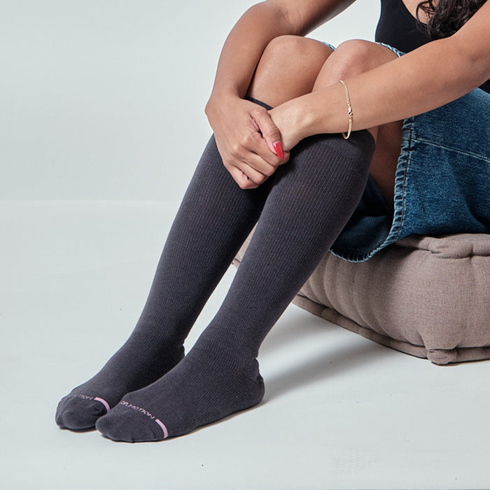 Knee-high compression socks in dark gray, ribbed texture, worn by a seated woman with crossed legs.