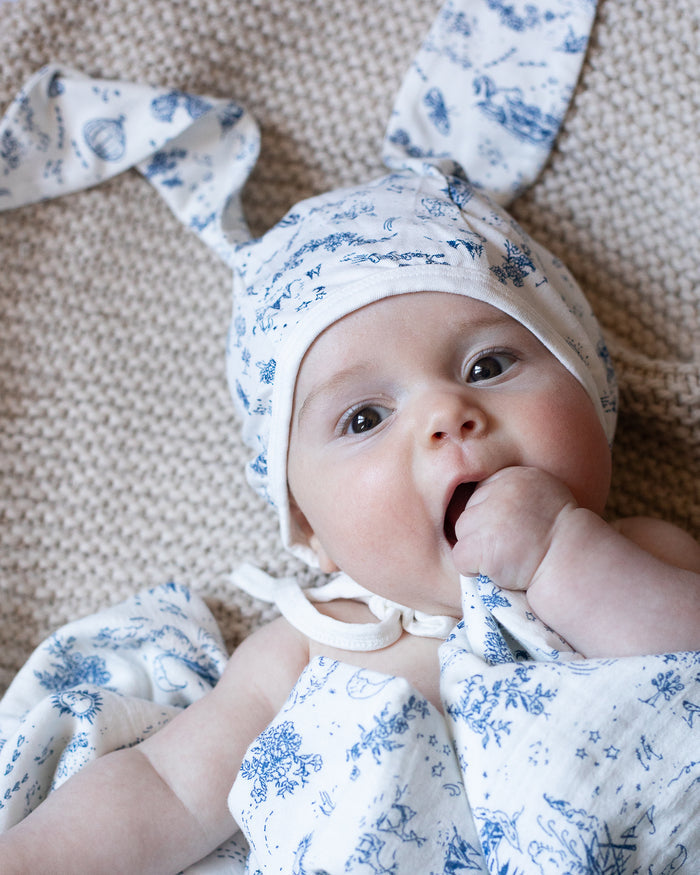 A baby wearing a blue and white floral bonnet with bunny ears, and wrapped in a blue-and-white toile blanket.
